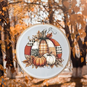 Può includere: Un telaio da ricamo a punto croce con un design a patchwork di zucca. La zucca è composta da vari motivi e colori, tra cui rosso, arancione e scozzese. Due zucche più piccole e foglie autunnali completano l'opera a tema autunnale.