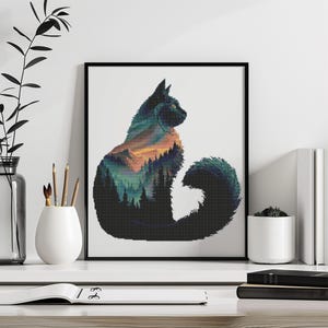 May include: Black framed art print featuring a cat silhouette with a landscape design. The cat's form is filled with a colorful mountain and forest scene, with shades of green, orange, and blue. The artwork is displayed on a white surface.