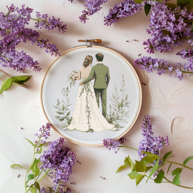 May include: A cross-stitch embroidery hoop with a design of a bride and groom standing together in a field of greenery. The bride is wearing a white dress and the groom is wearing a green suit.