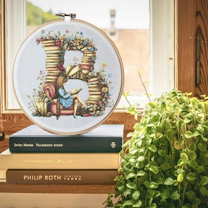 Op de afbeelding: Een witte borduurring met een kruissteekontwerp van de letter B gemaakt van gestapelde boeken. Een klein figuurtje zit op een stoel en leest een boek. De ring ligt op een stapel boeken met de titels "Javier Marias", "Valkoinen Sydan", "Tanssikon Emile", "PHILIP ROTH" en "Immisen Tahra".