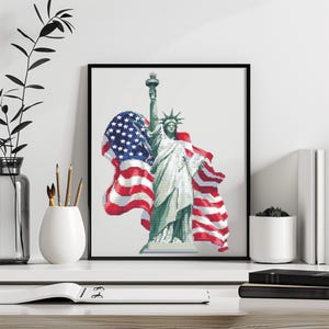 May include: Framed cross-stitch art featuring the Statue of Liberty with an American flag. The artwork is in shades of green, white, red, and blue, and is displayed on a white surface.