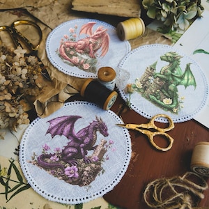 Op de afbeelding: Drie ronde gestikte stoffen patches met draken in verschillende kleuren. De roze draak is op een witte achtergrond met roze bloemen. De groene draak is op een witte achtergrond met groene bloemen. De paarse draak is op een witte achtergrond met paarse bloemen.