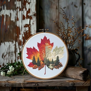 Peut inclure: Broderie au point de croix d'une feuille d'érable dans un cercle en bois. La feuille est divisée en sections rouges, oranges et vertes, avec une scène de forêt brodée au centre. La broderie est exposée sur une surface en bois rustique.