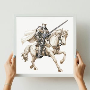 May include: A framed cross-stitch depicting a knight in full armor on a white horse. The knight holds a sword, and the horse is adorned with decorative elements. The artwork features a white background and is held by hands with blue nail polish.