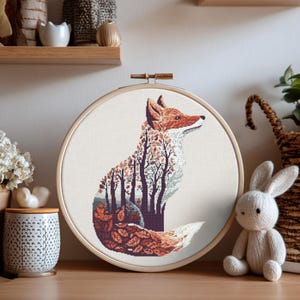 May include: A cross-stitch embroidery hoop with a fox design. The fox is depicted in orange, brown, and white, with a forest scene within its outline. The embroidery is set in a wooden hoop, creating a rustic aesthetic, suitable for home decor.