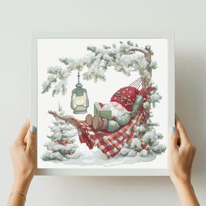 May include: Cross-stitch art of a gnome reading in a hammock. The gnome wears a red hat and rests on a red and white plaid blanket. A lantern hangs from a snow-covered tree branch. The scene is set in a snowy winter landscape.