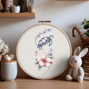 Könnte beinhalten: Ein runder Holzstickrahmen mit einem Kreuzstichmotiv auf weißem Stoff. Das Muster zeigt eine hellviolette Meeresschildkröte, die über einer großen rosa Hibiskusblüte schwimmt, akzentuiert durch kleine violette Blüten, grüne Blätter und Herzen.