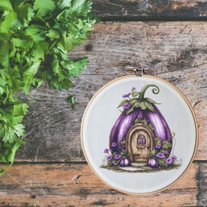 May include: A purple eggplant-shaped house with a brown door and a small window. The house is surrounded by green leaves and purple flowers. The house is embroidered on a white fabric and framed in a wooden hoop.