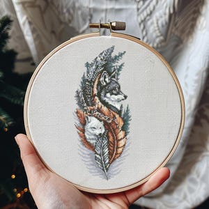 Pode incluir: Um bastidor de bordado em ponto cruz com um design detalhado de dois lobos, um cinzento e outro branco, rodeados por penas e folhagens. A obra de arte é definida contra um tecido creme, preso dentro de um aro de madeira.