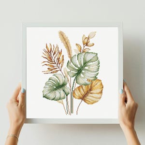 May include: A framed cross-stitch artwork featuring a botanical arrangement of tropical leaves and grasses in shades of green, gold, and brown. The artwork is held by a person with light blue nail polish.