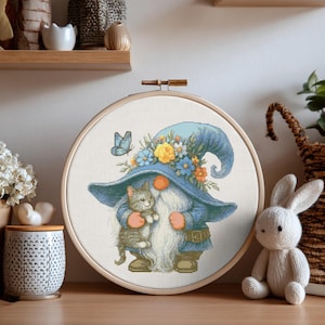 Peut inclure: Broderie au point de croix dans un cercle en bois. Le motif représente un gnome avec un chapeau et un manteau bleus, tenant un chat tabby gris. Le chapeau est orné de fleurs et d'un papillon. Le fond est blanc.
