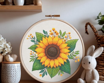 Puede incluir: Un bordado de punto de cruz de un girasol en un aro de madera. El girasol tiene pétalos amarillos y un centro marrón oscuro, rodeado de hojas verdes y detalles amarillos. El bordado se exhibe sobre una superficie de madera.