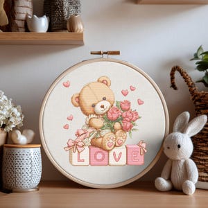 Puede incluir: Un bastidor de bordado de punto de cruz con un oso de peluche sosteniendo rosas rosas y un lazo rosa. La palabra "LOVE" está escrita con letras rosas. Corazones rosas están esparcidos alrededor del oso. El bastidor está sobre un fondo neutro.