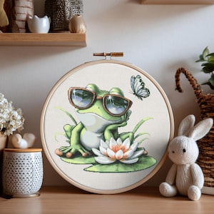 May include: A cross-stitch embroidery hoop showing a green frog in sunglasses, relaxing on a lily pad with a white and orange flower. A butterfly is also present. The hoop is displayed on a wooden surface.