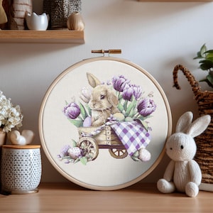 May include: A cross-stitch embroidery in a wooden hoop features a bunny in a wooden cart filled with purple tulips and Easter eggs. A purple and white checkered blanket covers the cart. A stuffed bunny sits nearby.