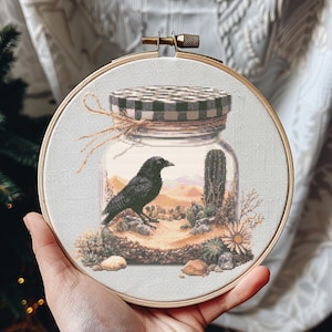 Op de afbeelding: Een zwarte raaf zit in een glazen pot met een geruit deksel. De pot bevat een woestijnscène met cactussen en zand.