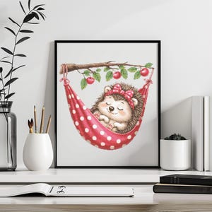 Könnte beinhalten: Gerahmter Druck mit einer Illustration eines schlafenden Igels in einer roten Polka-Dot-Hängematte. Der Igel trägt eine rote Schleife und ruht unter einem Ast mit Kirschen. Das Kunstwerk wird auf einer weißen Oberfläche mit anderen Objekten im Vordergrund gezeigt.