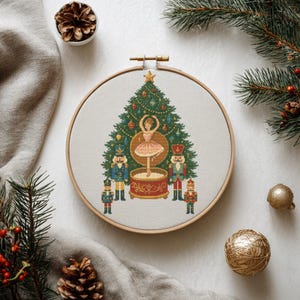 May include: A cross-stitch embroidery hoop featuring a Christmas tree design with a ballerina and nutcrackers. The tree is decorated with ornaments. The hoop is made of wood and is surrounded by pine needles and gold baubles.