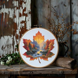 Pode incluir: Bordado em ponto cruz num bastidor de madeira, com um desenho de folha de bordo. A folha é preenchida com uma paisagem de árvores de outono, um rio e um pôr do sol. As cores incluem tons de laranja, amarelo, verde e azul.