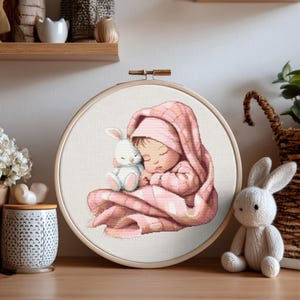 Può includere: Un telaio da ricamo a punto croce con un bambino che dorme avvolto in una coperta e un cappello rosa, che tiene in braccio un coniglietto bianco. Il disegno è su sfondo bianco, incorniciato da un cerchio di legno.