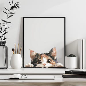 Könnte beinhalten: Ein gerahmter Kunstdruck mit einer neugierigen Schildpattkatze, die über eine Oberfläche blickt. Das Gesicht der Katze ist detailliert mit braunem, orangefarbenem und weißem Fell, großen Augen und einer rosa Nase. Das Kunstwerk ist vor einem weißen Hintergrund.