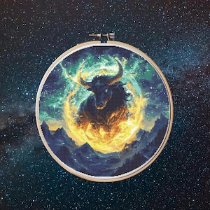 May include: A cross-stitch embroidery hoop with a celestial bull design. The bull is dark blue with fiery orange and yellow accents, set against a starry night sky. The artwork is framed in a wooden hoop, a common craft item.