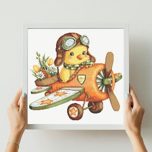Può includere: Uno schema a punto croce incorniciato raffigurante un pulcino giallo con un cappello da aviatore marrone e occhiali protettivi, seduto su un aereo arancione. L'aereo è decorato con stelle arancioni sulle ali e sulle ruote e porta dei fiori.