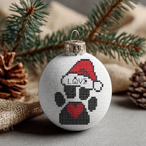 May include: White Christmas ornament with a cross-stitch design featuring a black paw print wearing a red Santa hat. The word "LOVE" is stitched above the paw print, and a red heart is in the center of the paw print.