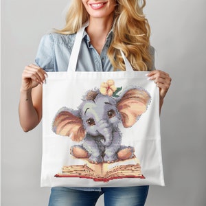 Pode incluir: Sacola de tecido branca com um elefante de desenho animado fofo usando uma flor na cabeça e sentado em um livro vermelho. O elefante é cinza com orelhas cor-de-rosa e uma tromba azul claro.
