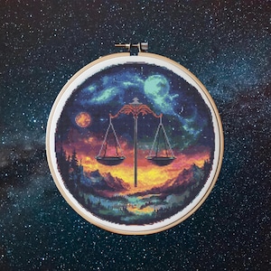 May include: A round embroidery hoop featuring a colourful cross-stitch design of a Libra zodiac symbol. The design depicts a scale set against a starry night sky with a landscape of mountains and a river.