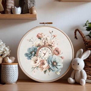 Könnte beinhalten: Kreuzstich-Stickerei in einem Holzreifen. Das Design zeigt eine Taschenuhr, umgeben von pastellfarbenen Blumen in Rosa, Blau und Creme. Die Zeiger der Uhr zeigen auf etwa 10:10 Uhr. Der Reifen wird auf einer Holzoberfläche präsentiert.