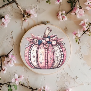 May include: A cross-stitch pumpkin design in a wooden embroidery hoop. The pumpkin is primarily pink with white and darker pink accents, adorned with a bow and embellishments. The background is a neutral color, with pink flowers surrounding the hoop.