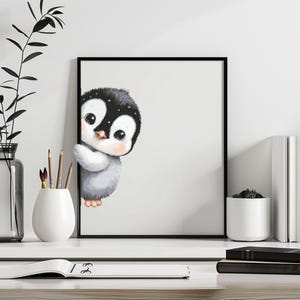 May include: A framed print featuring a cute cartoon penguin with a black and white head, gray body, and large eyes. The penguin is peeking out from behind a white background. The artwork is displayed on a white shelf with decorative items.