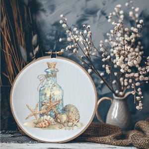 May include: A blue glass bottle with a cork stopper filled with seashells and starfish, surrounded by additional seashells on a white background. The bottle and seashells are in a round wooden embroidery hoop.