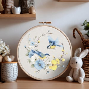 May include: A cross-stitch embroidery hoop featuring a design of two blue birds surrounded by blue, white, and yellow flowers. The birds have yellow bellies and blue wings. The embroidery is set in a wooden hoop.