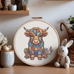May include: A circular wooden embroidery hoop containing a cross-stitch pattern of a seated calf. The calf features a colorful patchwork design with various geometric patterns in shades of blue, orange, purple, and yellow on a white fabric background.