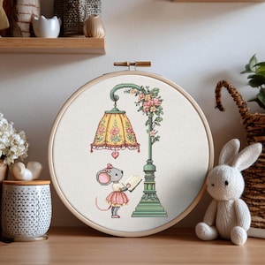 Puede incluir: Un bordado de punto de cruz en un bastidor de madera redondo que muestra un ratón gris con falda y lazo rosa, leyendo un libro abierto bajo una farola verde. La pantalla amarilla con motivos florales está decorada con rosas y un corazón.