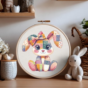 May include: A cross-stitch embroidery of a patchwork bunny in a wooden hoop. The bunny has a large bow, long ears, and is seated. The color palette includes pink, blue, yellow, and green. A small stuffed bunny sits nearby.