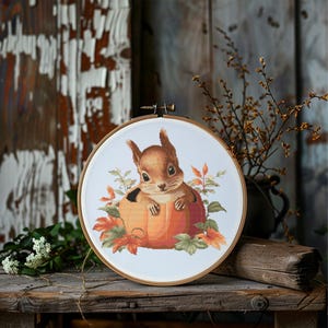 May include: A cross-stitch embroidery hoop featuring a brown squirrel nestled in an orange pumpkin, surrounded by fall leaves. The artwork is set against a rustic wooden background, creating a cozy, autumnal theme.