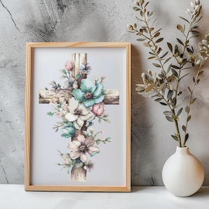 May include: A framed cross-stitch artwork depicting a wooden cross decorated with pastel-coloured flowers. The flowers feature shades of pink, white, and teal, creating a soft, floral design. The artwork is displayed in a light-coloured wooden frame.
