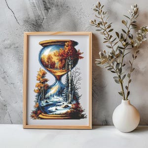 Può includere: Una stampa incorniciata con un design a clessidra pixelato. La clessidra contiene una scena paesaggistica che mostra l'autunno e l'inverno. La cornice è in legno chiaro. L'opera d'arte è esposta su una superficie bianca, con un vaso e del fogliame.