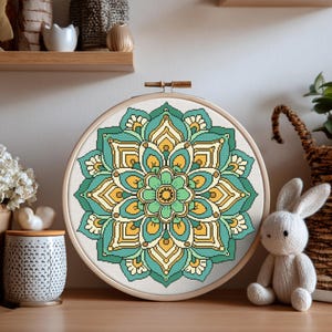 May include: A completed cross-stitch mandala design in a wooden hoop. The mandala features a symmetrical pattern in shades of teal, yellow, and green. The embroidery is displayed on a wooden surface, with a decorative ceramic jar and a stuffed bunny in the background.