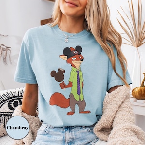 Puede incluir: Camiseta azul claro con un personaje de zorro de dibujos animados que lleva un sombrero de Mickey Mouse y sostiene un helado de Mickey Mouse. El zorro viste una camisa verde, una corbata morada y pantalones marrones. La palabra "Chambray" est&aacute; impresa en la parte inferior izquierda.
