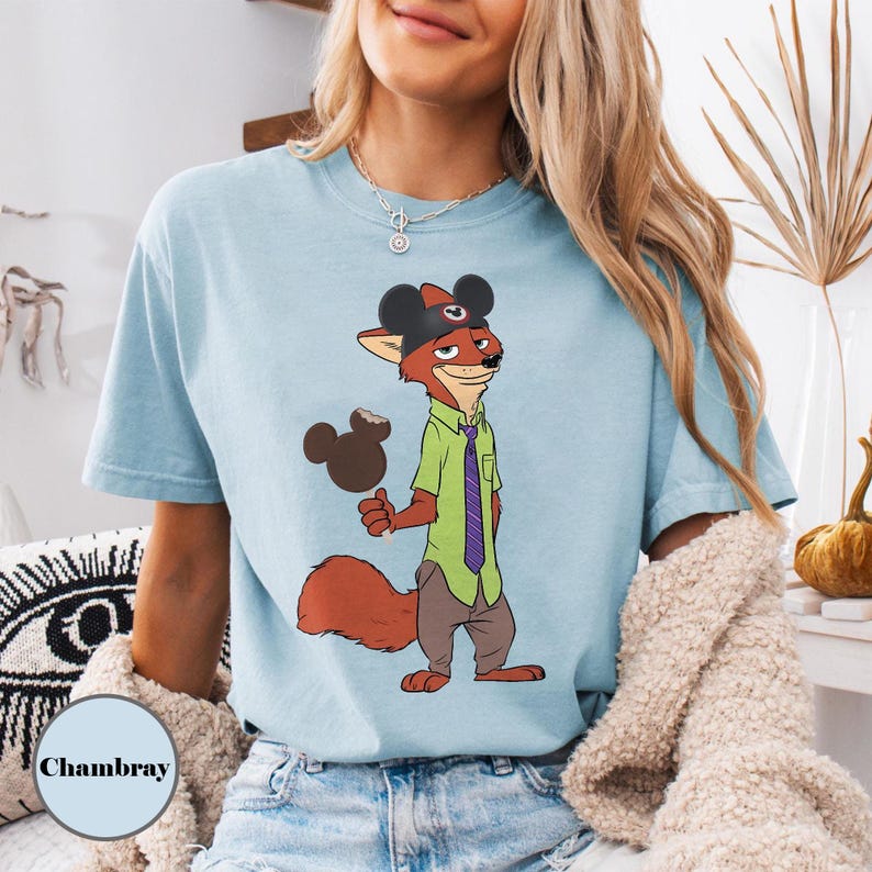 Puede incluir: Camiseta azul claro con un personaje de zorro de dibujos animados que lleva un sombrero de Mickey Mouse y sostiene un helado de Mickey Mouse. El zorro viste una camisa verde, una corbata morada y pantalones marrones. La palabra "Chambray" est&aacute; impresa en la parte inferior izquierda.