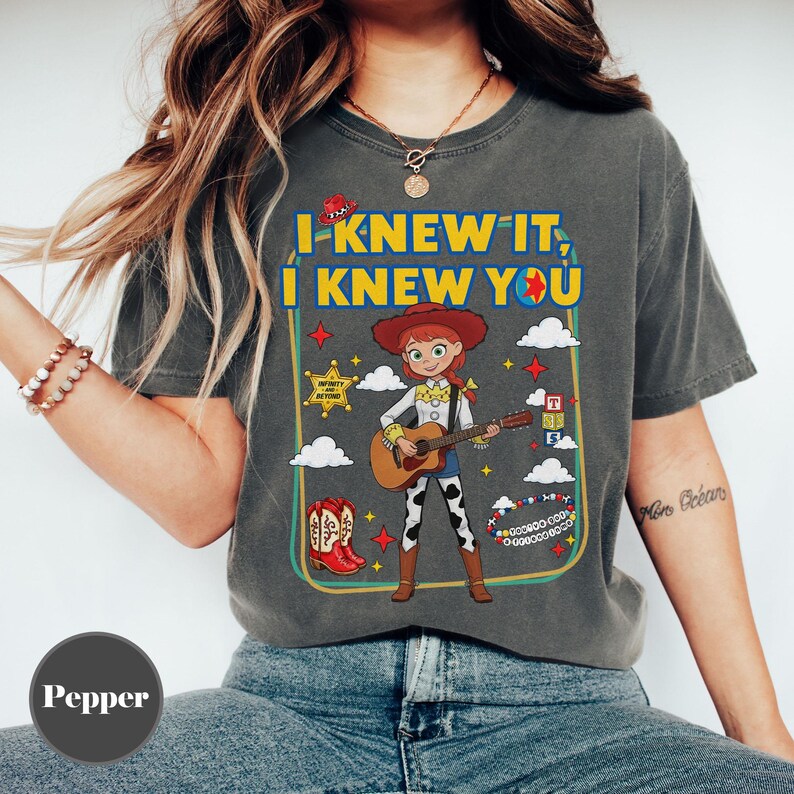 May include: A charcoal gray t-shirt featuring a graphic of a cowgirl character playing an acoustic guitar. The text reads I KNEW IT, I KNEW YOU. The design includes a sheriff star, red cowboy boots, alphabet blocks, and a beaded bracelet.