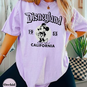 May include: A light purple t-shirt featuring the Disneyland logo in black script above a graphic of Minnie Mouse. The graphic includes the numbers "19" and "55" and the word "CALIFORNIA". The shirt is a casual style.