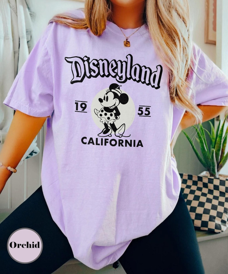 May include: A light purple t-shirt featuring the Disneyland logo in black script above a graphic of Minnie Mouse. The graphic includes the numbers "19" and "55" and the word "CALIFORNIA". The shirt is a casual style.