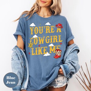 Puede incluir: Una camiseta azul de manga corta con el texto amarillo "YOU'RE A COWGIRL LIKE ME", que incluye gr&aacute;ficos de un sombrero de vaquero rojo, una estrella de sheriff amarilla y botas rojas, decorada con nubes blancas y peque&ntilde;as estrellas rojas y amarillas.