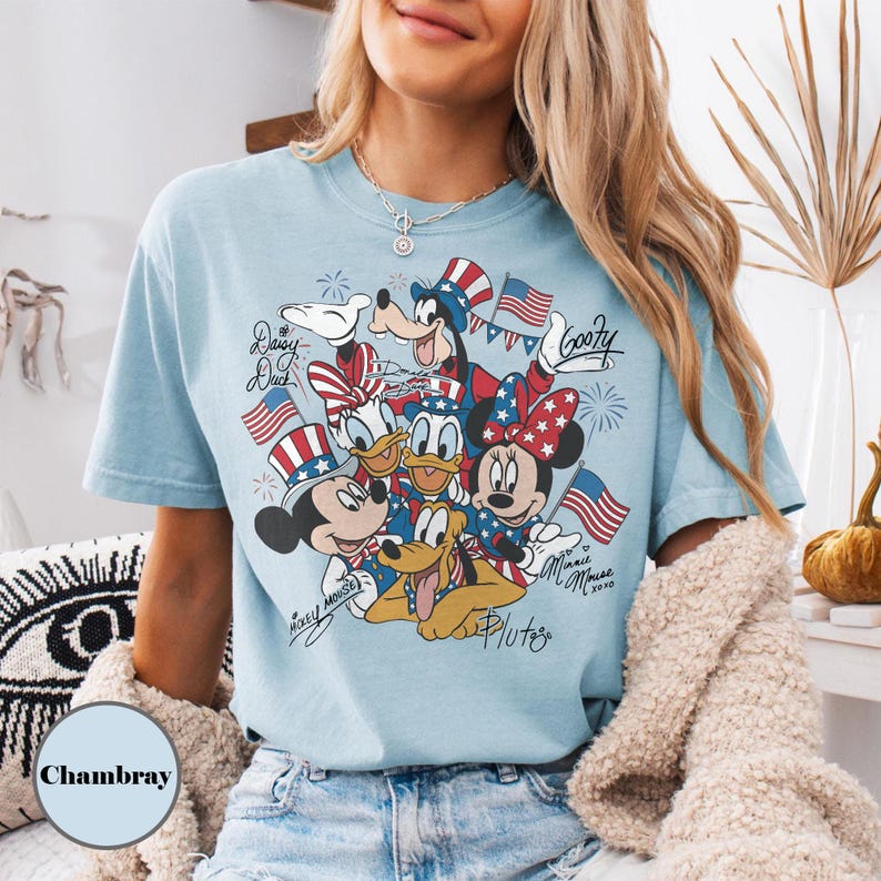 Peut inclure: T-shirt bleu clair avec un motif de personnages Disney, Mickey Mouse, Minnie Mouse, Donald Duck, Daisy Duck, Dingo et Pluto, avec des drapeaux am&eacute;ricains et des accents patriotiques. Le t-shirt porte l'inscription "Minnie Mouse xoxo" et "Mickey Mouse".