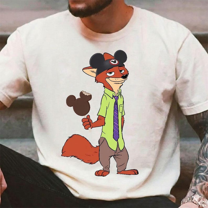 Puede incluir: Camiseta color crema con un personaje de zorro de dibujos animados que lleva un sombrero de Mickey Mouse y sostiene un helado con forma de Mickey Mouse. El zorro viste una camisa verde, una corbata morada y pantalones marrones. El dise&ntilde;o es un gui&ntilde;o juguet&oacute;n a Disney.